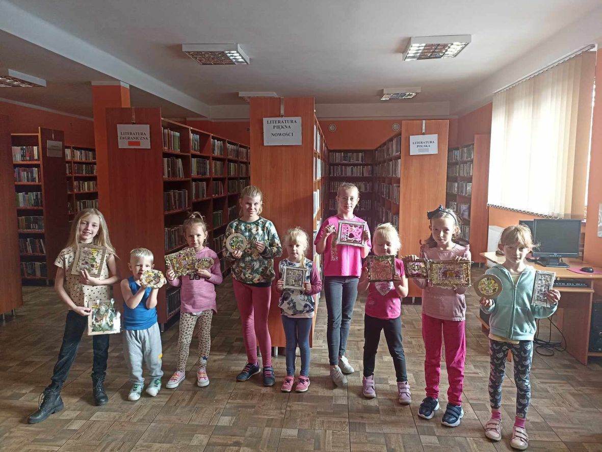 Dziewięcioro dzieci w bibliotece, prezentują jesienne witraże wykonane samodzielnie, w tle regały z książkami.