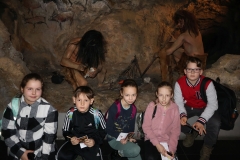 zdjęcie uczestników półkolonii. Za siedzącymi dziećmi widać ekspozycję przedstawiającą człowieka pierwotnego odkrywającego ogień. 