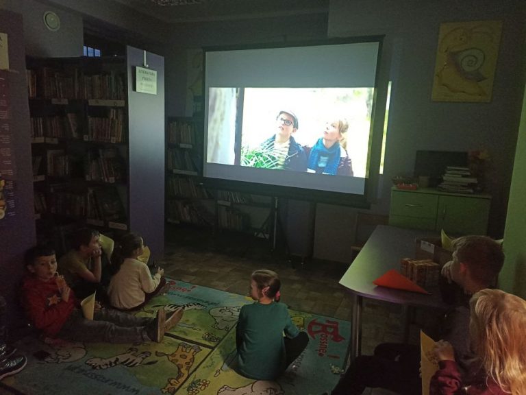 Sala biblioteczna, dzieci na dywanie oglądają film w bibliotece i jedzą popcorn.