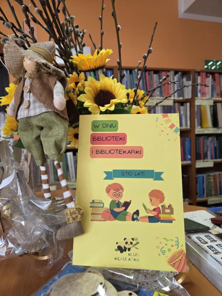 Kartka z życzeniami na tydzień bibliotek, na żółtym tle siedzi dwoje dzieci i czyta książki. tekst z kartki W dniu biblioteki i bibliotekarki Sto Lat!
