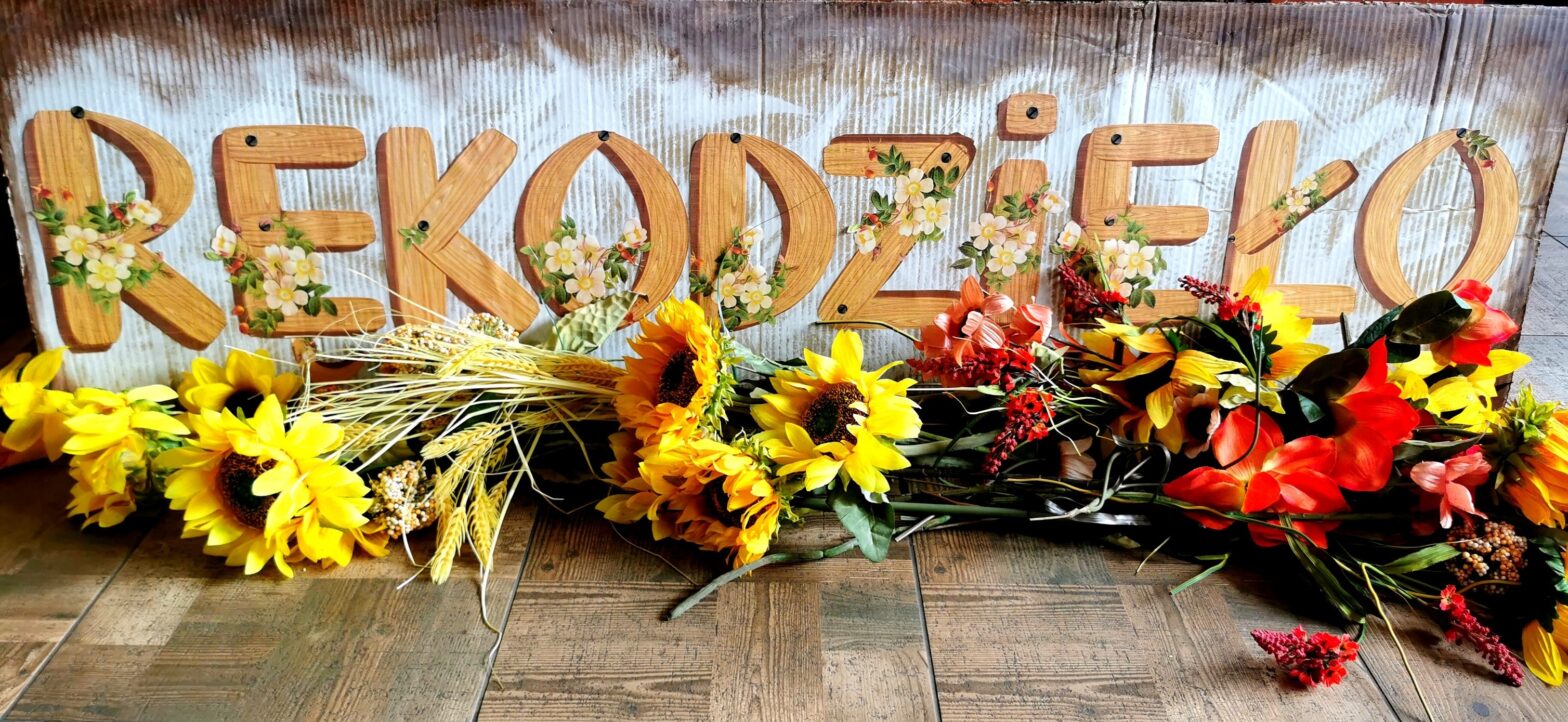 Napis na kartonie "rękodzieło", poniżej kwiaty polne takie jak żółte słoneczniki i kłosy zboża.