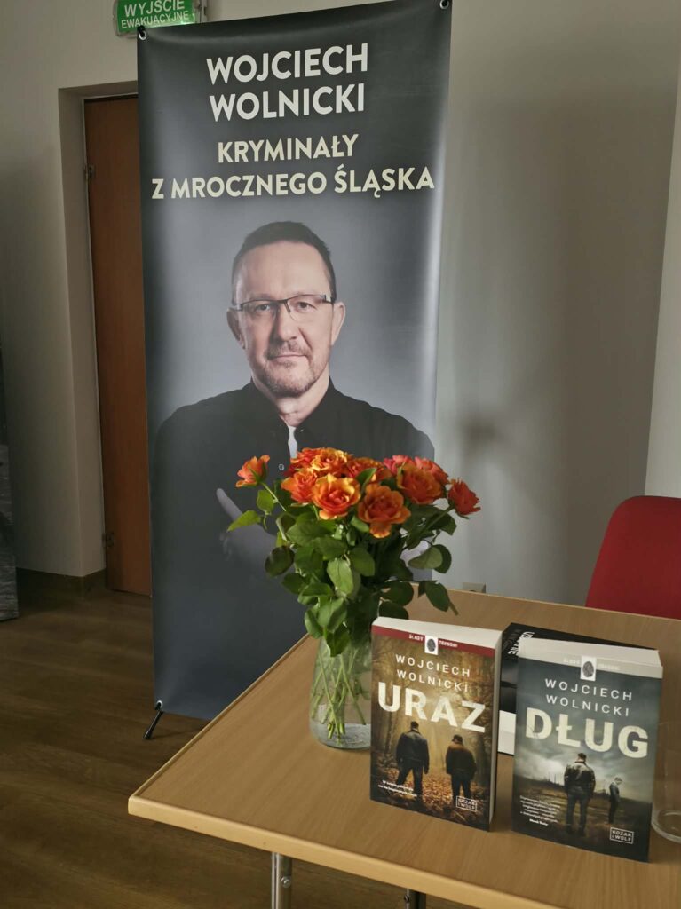 Na zdjęciu stolik z kwiatami i dwie książki w tle baner ze zdjeciem wojciecha wolnickiego z napisem kryminały z mrocznego śląska