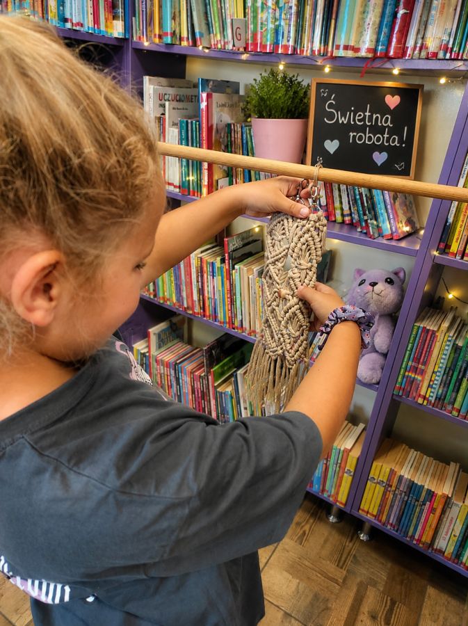 Na zdjęciu widać dziecko podczas zajęć plastyczno-rękodzielniczych w bibliotece. Dziewczynka trzyma własnoręcznie wykonany, beżowy brelok z makramy — ozdobę z misternie splecionych sznurków, metalowym karabińczykiem i długimi frędzlami. W tle znajdują się kolorowe książki na fioletowych regałach, a ciepłe światełka, roślina w różowej doniczce, pluszowa zabawka i tabliczka z napisem „Świetna robota!” tworzą przyjazną, radosną atmosferę zajęć.