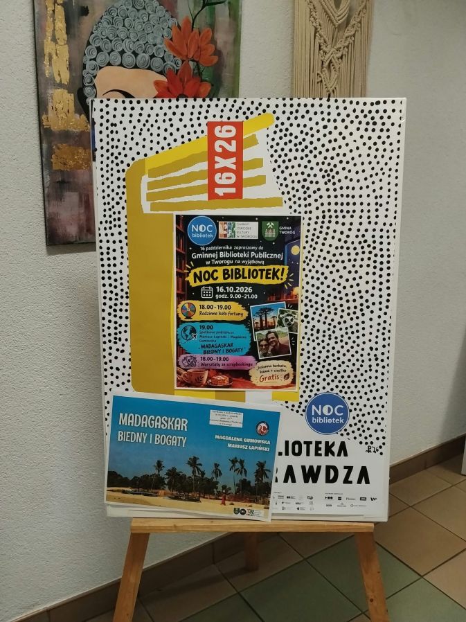 Pionowy plakat reklamowy zapowiadający wydarzenie „Noc Bibliotek 2026”, umieszczony na drewnianej sztaludze w jasnym pomieszczeniu. Tło plakatu jest białe, gęsto pokryte drobnymi, czarnymi kropkami. Głównym elementem graficznym jest duży, żółty kształt przypominający ułożone w stos książki, na którym umieszczono ciemnoniebieską, prostokątną grafikę szczegółowo opisującą program wydarzenia. Na samej górze żółtego kształtu widnieje pionowy, pomarańczowy prostokąt z białym napisem „16. X. 26” (oznaczającym datę 16 października 2026). W dolnej części plakatu, po lewej stronie, przypięta jest mniejsza ulotka ze zdjęciem egzotycznej plaży z palmami, promująca spotkanie podróżnicze pt. „Madagaskar. Biedni i bogaci” autorstwa Magdaleny Gumowskiej i Mariusza Łapińskiego. Po prawej stronie na dole widoczny jest fragment dużego, czarnego napisu „BIBLIOTEKA SIĘ SPRAWDZA” oraz logo akcji „Noc Bibliotek”. W tle, za sztalugą, widać fragment ściany z nowoczesnym, kolorowym obrazem przedstawiającym twarz buddy z czerwonymi kwiatami we włosach.